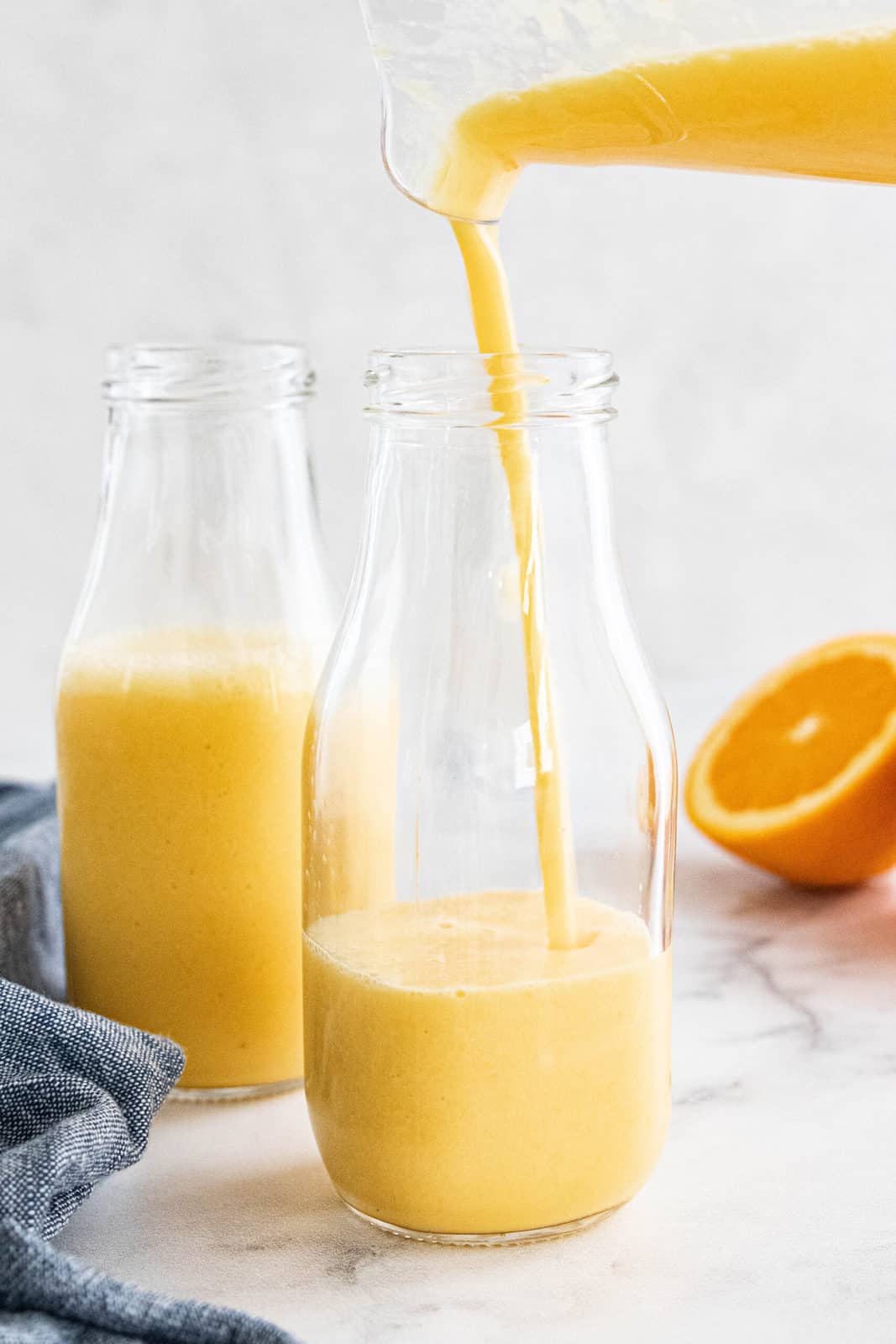 A pitcher pouring an orange smoothie into a glass jar that is in front of a full glass jar.