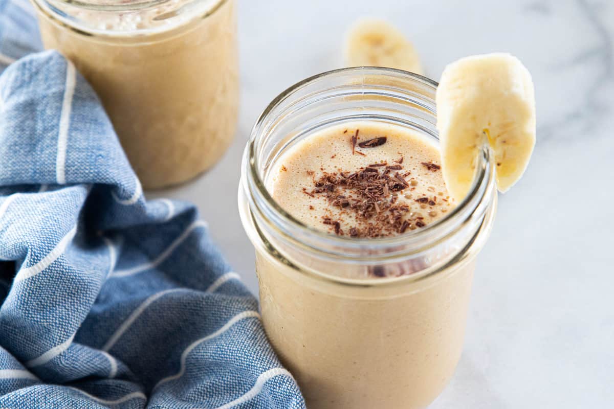 Two peanut butter powder smoothies in glasses with a slice of banana on the side of one glass. 