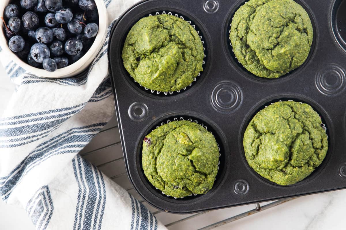 A muffin tin of cooked veggie muffins on a cooling rack next to a bowl of fresh blueberries and a dish towel all on top of a granite countertop.