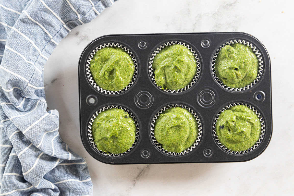 A muffin tin filled with spinach muffin batter next to a blue dish towel all on top of a granite countertop. 