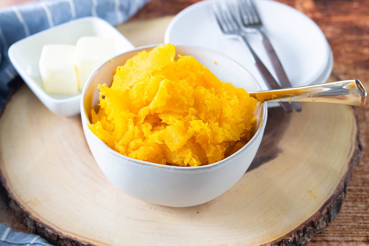 Steamed butternut squash in a white bowl with a dish of butter, two plates and two forks next to the squash all on top of a wood round.