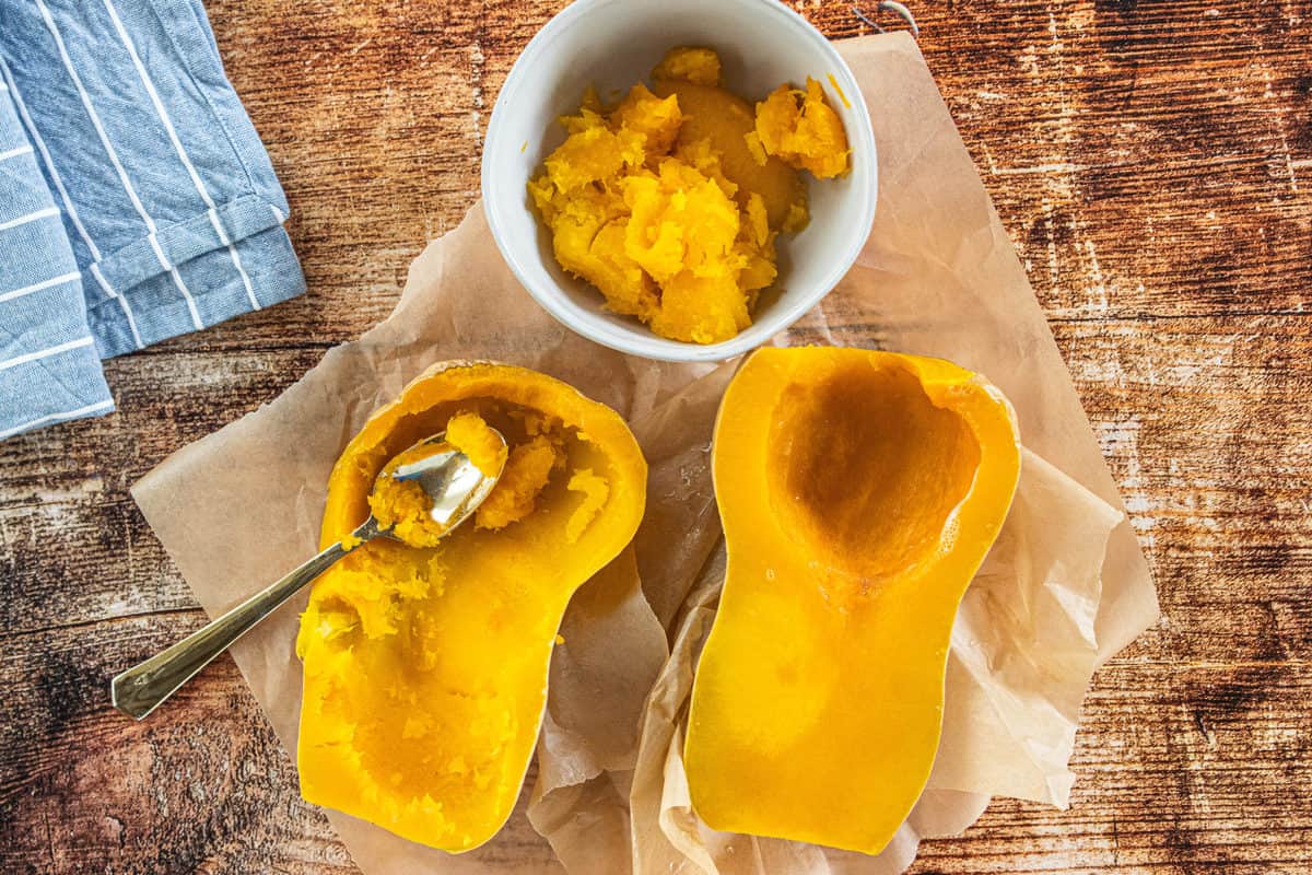 A microwave steamed butternut squash cut in half that is on top of brown parchment paper with a spoon that has scooped out squash into a white bowl.