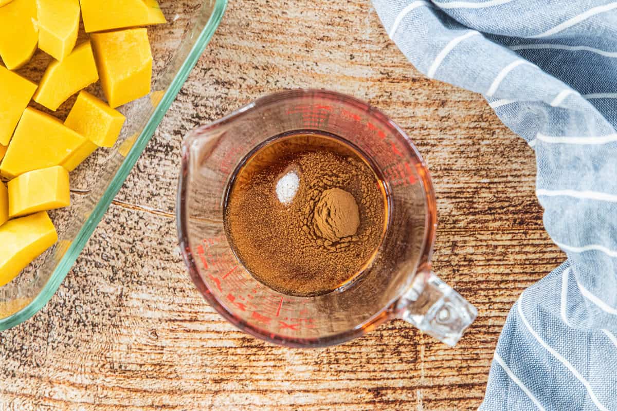 Ingredients in a liquid measuring cut next to chopped butternut squash in a glass pan and a dish towel all on top of a wood countertop.