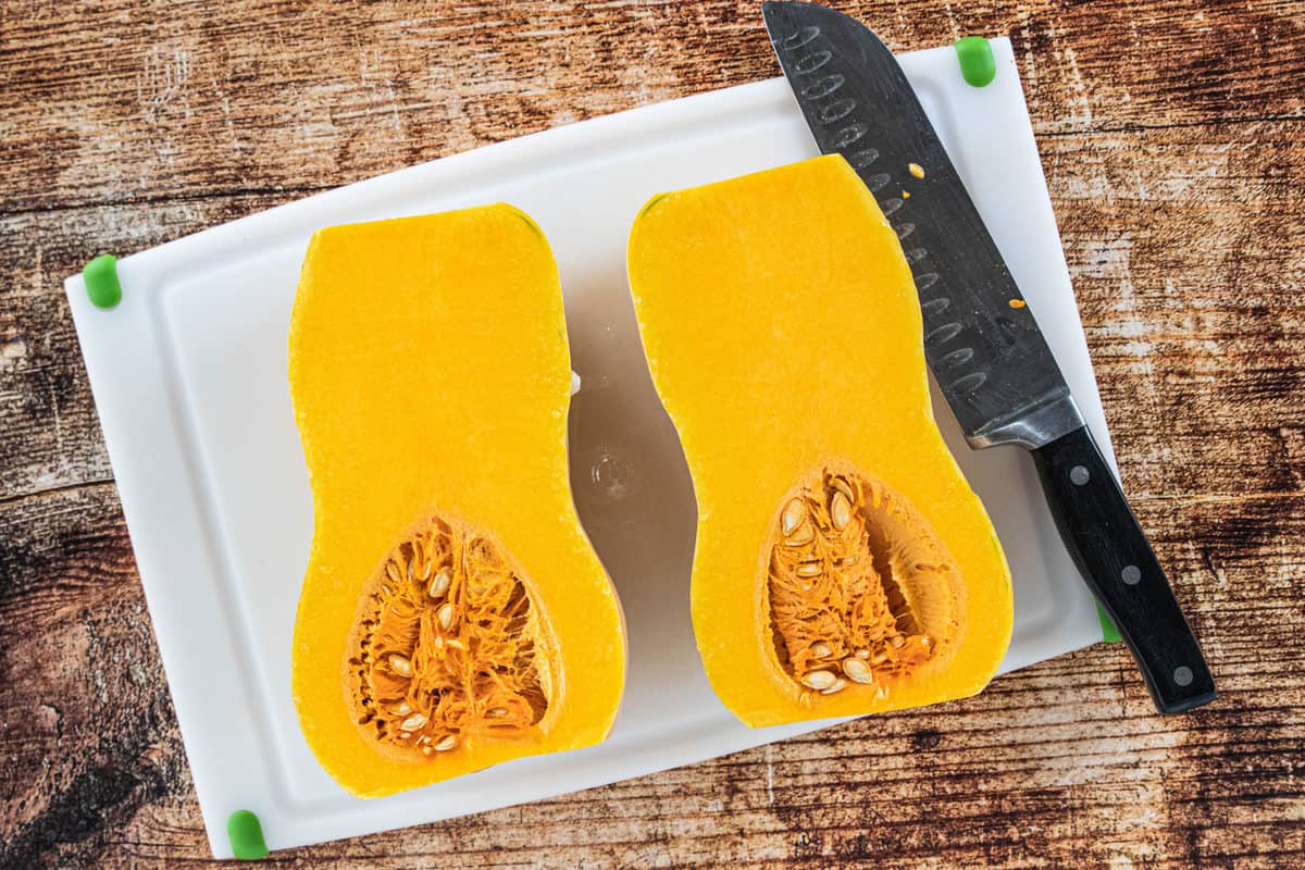 A butternut squash cut in half on top of a white cutting board with a knife on the cutting board all on top of a wood countertop. 