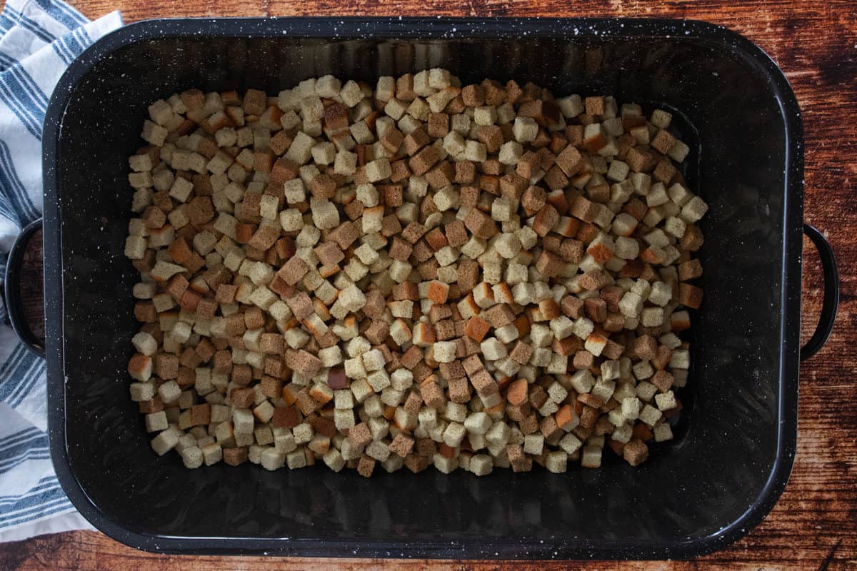 Bread cubes being shown in a large roasting pan that is sitting on top of a wooden countertop.