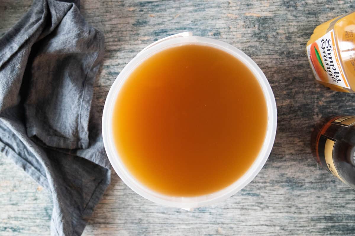 An ice cream pail filled with simple syrup, tea, lemonade, orange juice and brandy on top or a countertop. 