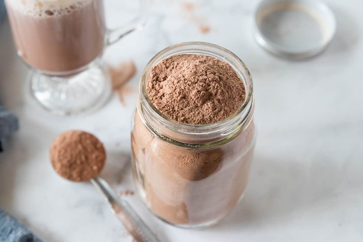 A mason jar filled with hot chocolate mix with a tablespoon full of mix next to the jar. A glass mug of hot chocolate sits behind the jar all on top of a countertop.
