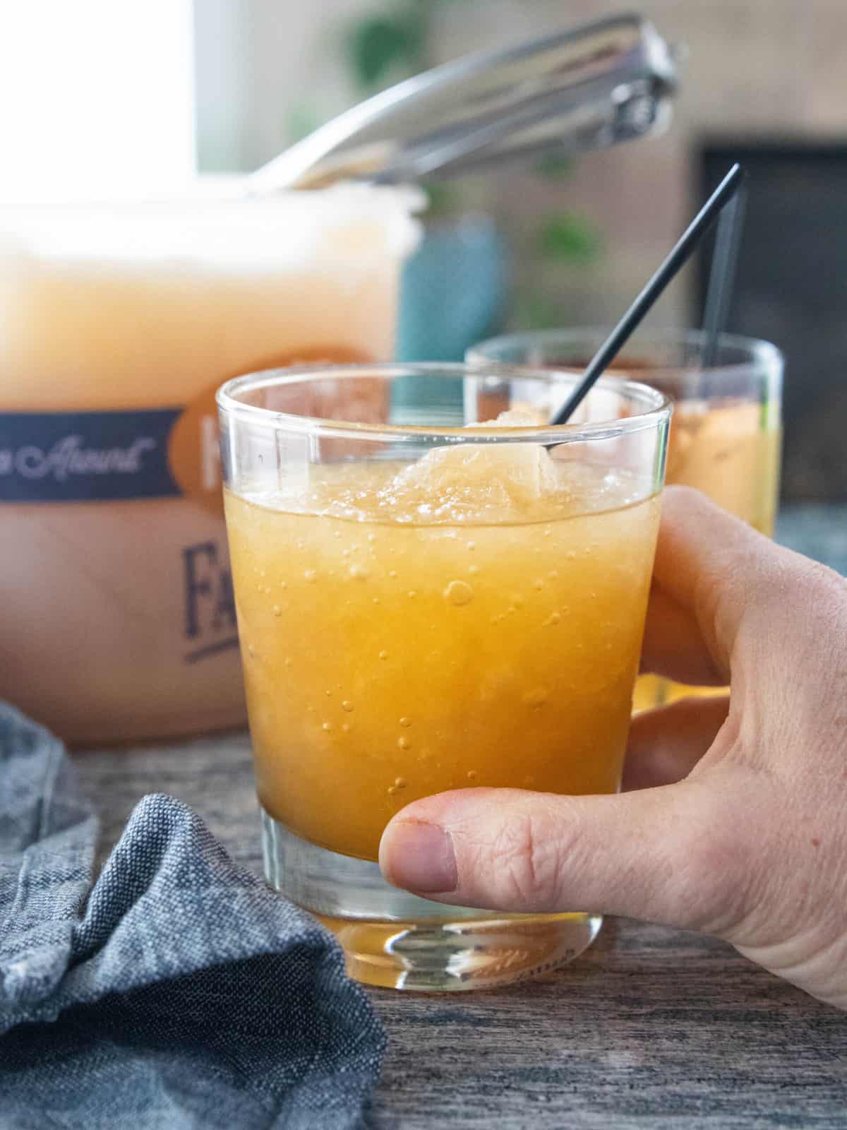 A hand holding a glass of brandy slush topped with soda with an ice cream pail of alcoholic slush behind the glass. 