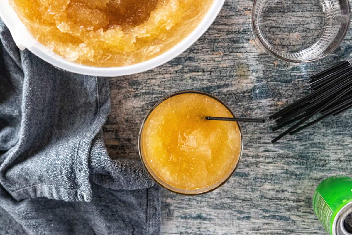 A glass filled with slush and a drink straw being shown with an ice cream pail filled with slush and a can of soda next to the glass all on top of a countertop.