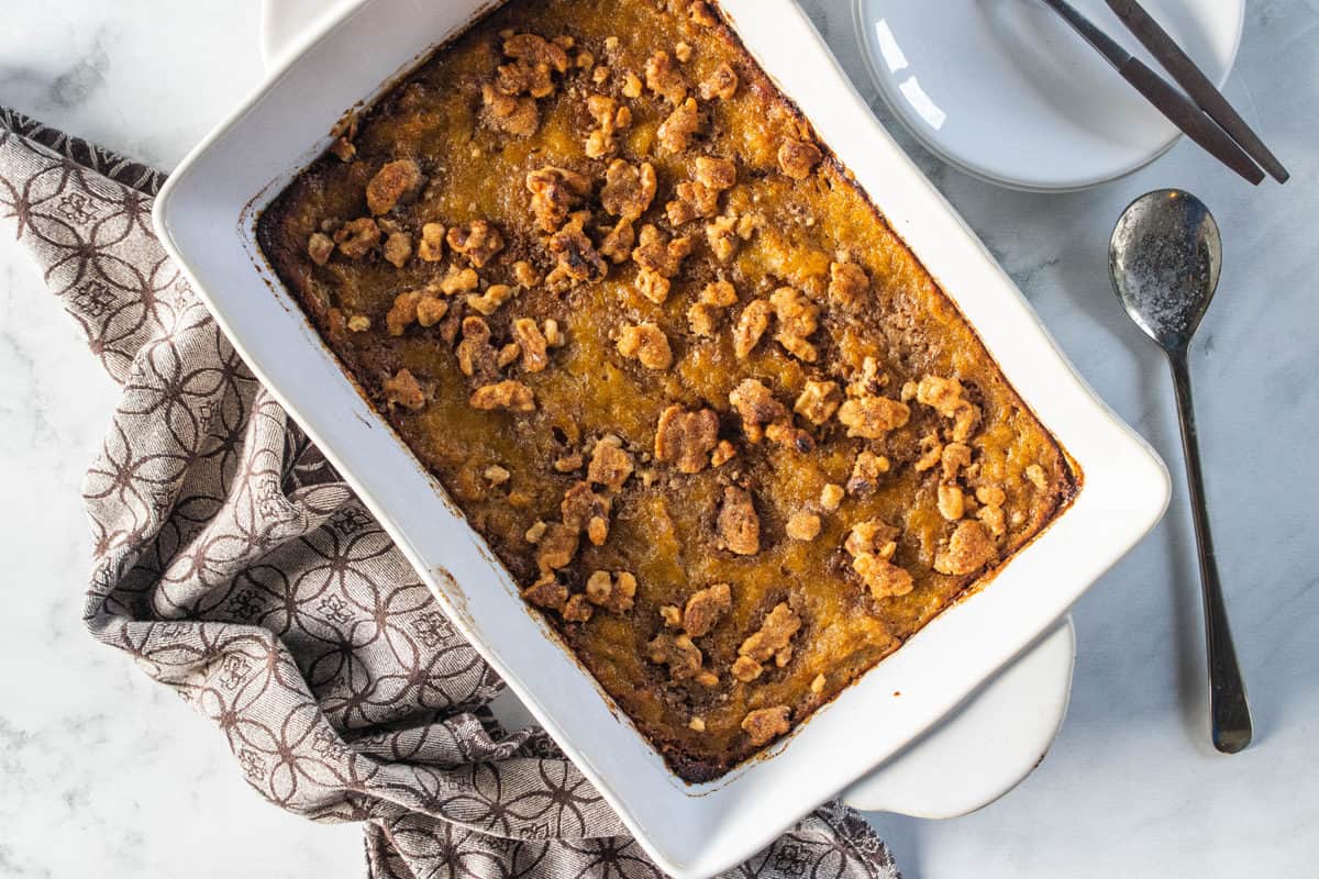 A top down view of a white pan with sweet potato casserole out of the oven next to a serving spoon and kitchen towel.