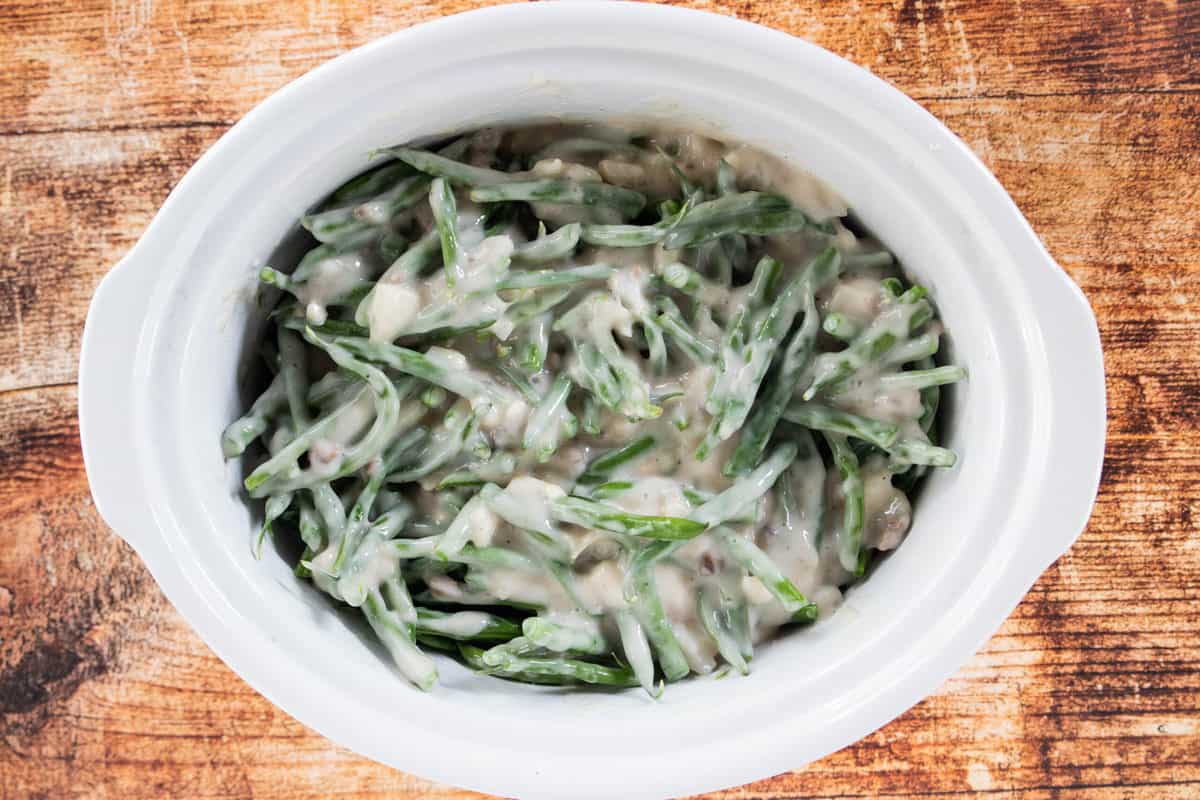 An uncooked casserole recipe being shown in a white crock pot that is on top of a wooden countertop.