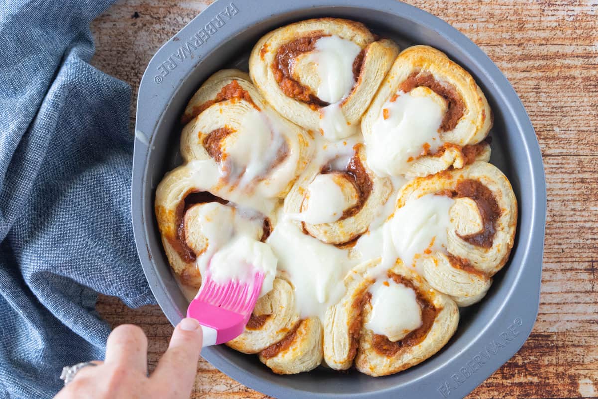 Sliced cinnamon rolls being brushed with icing while in a pie pan with a blue dish towel next to the pan all on top of a wood countertop.