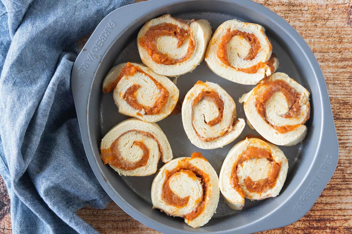 The roll sliced into cinnamon rolls and placed into a pie pan with a blue dish towel next to the pan all on top of a wood countertop.