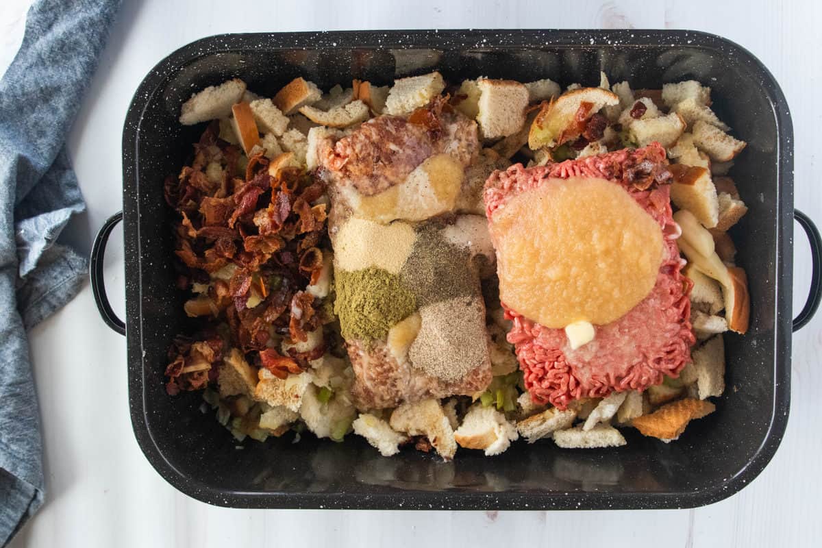 Pieces of torn white bread, bacon, spices, chopped celery, applesauce, sausage and ground beef in a large baking pan.