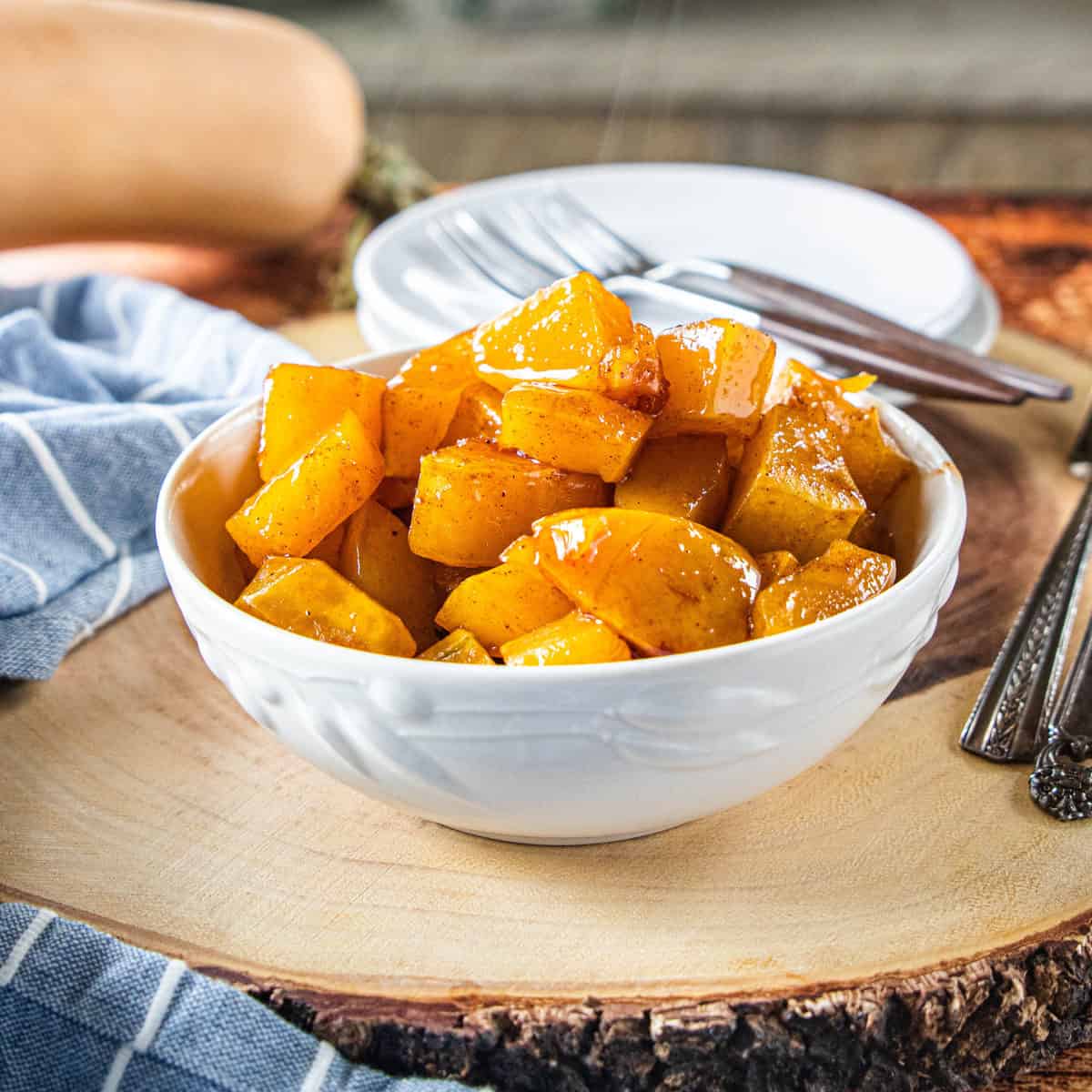 Maple roasted butternut squash in a white serving bowl on top of a wood plank with silverware and a dish towel around the bowl.