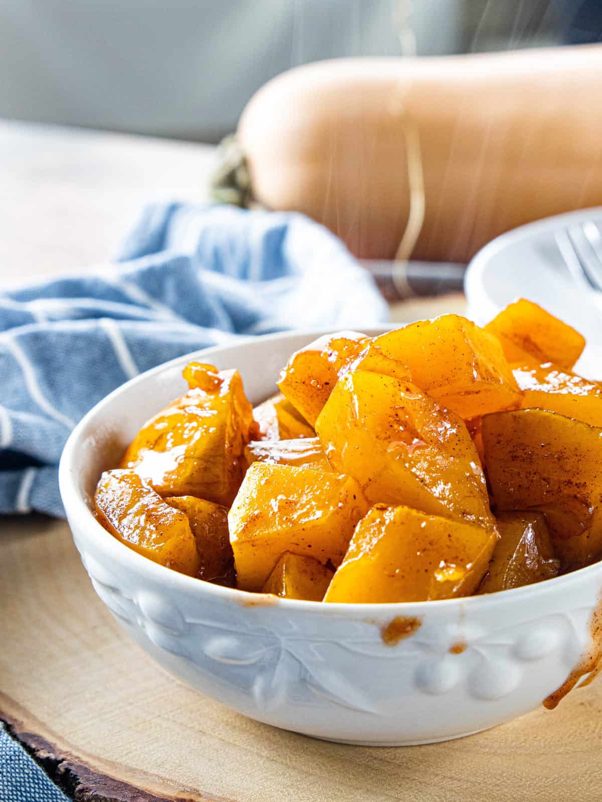 Roasted butternut squash in a white serving bowl on top of a wood plank with a dish towel next to the plank.