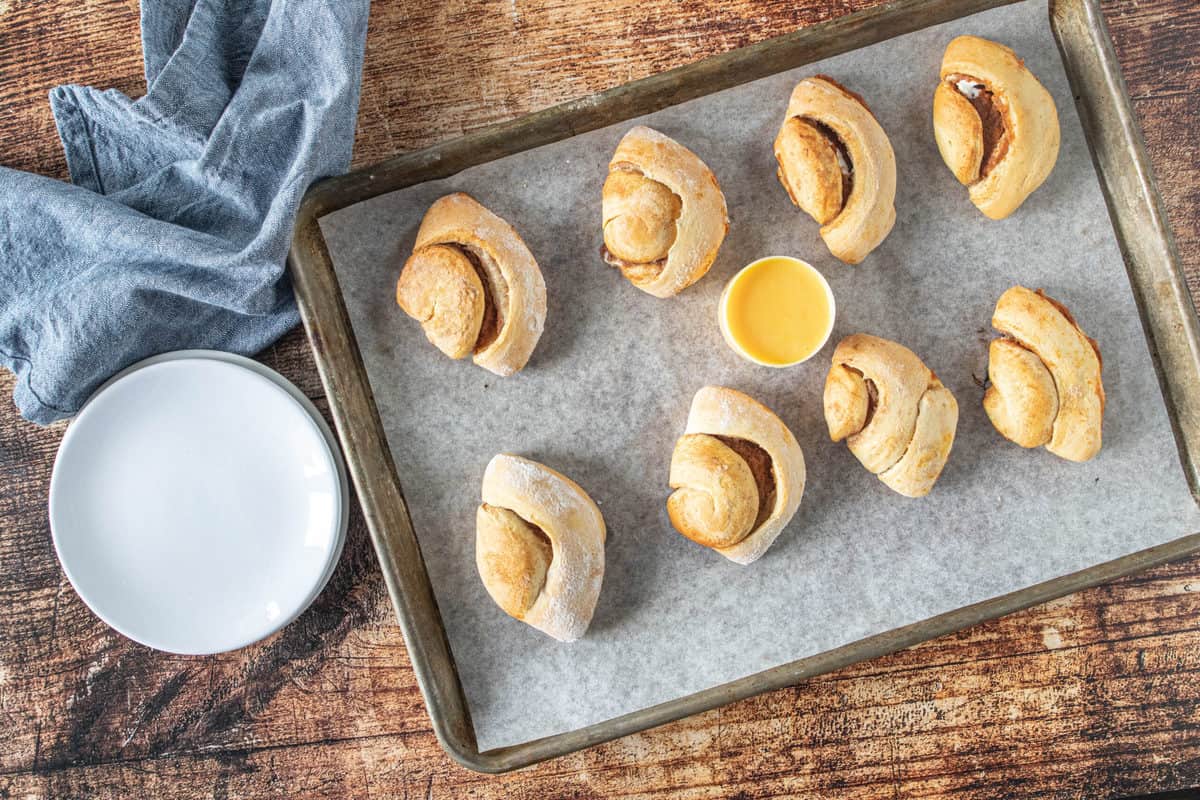 Baked pumpkin empanadas on a parchment lined baking sheet with a container of frosting in the middle of the baking sheet.
