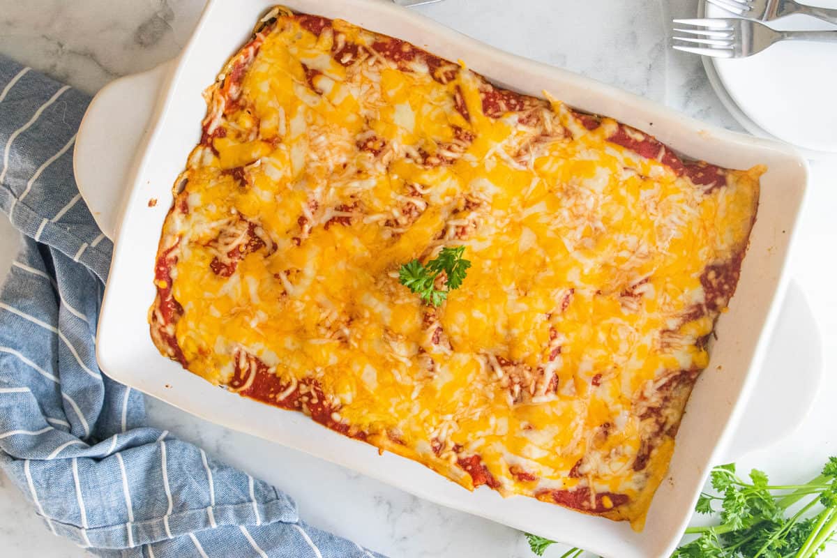 Baked spaghetti being shown in a white baking dish on top of a countertop with serving dishes and silverware next to the dish. 