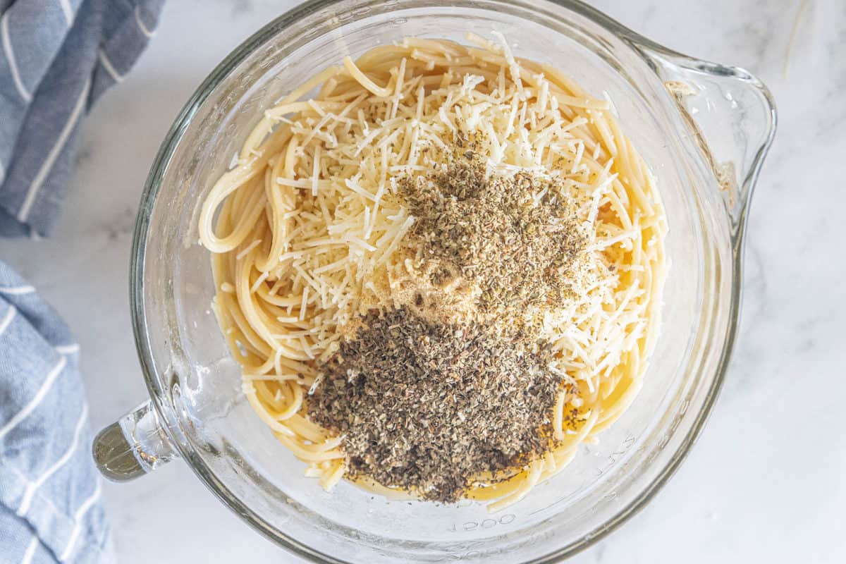 A mixing bowl being shown filled with cooked spaghetti, Spices, Parmesan cheese and an egg. 