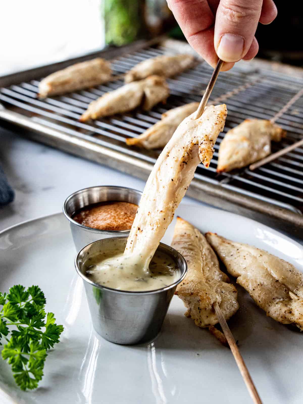 Chicken on a stick being shown dipped into a ramekin filled with a dipping sauce. 