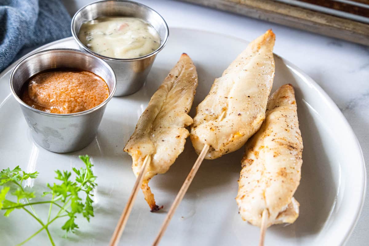 Chicken sticks being shown on a white plate with two ramekins on the plate filled with dipping sauces.