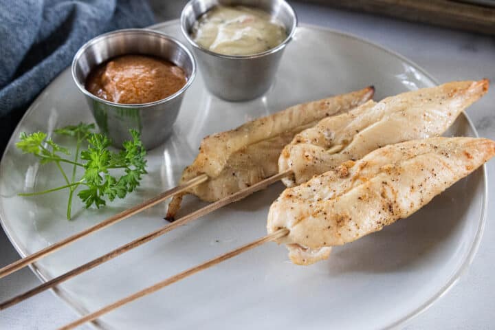 Chicken sticks being shown on a white plate with two ramekins filled with dipping sauces on the plate. 