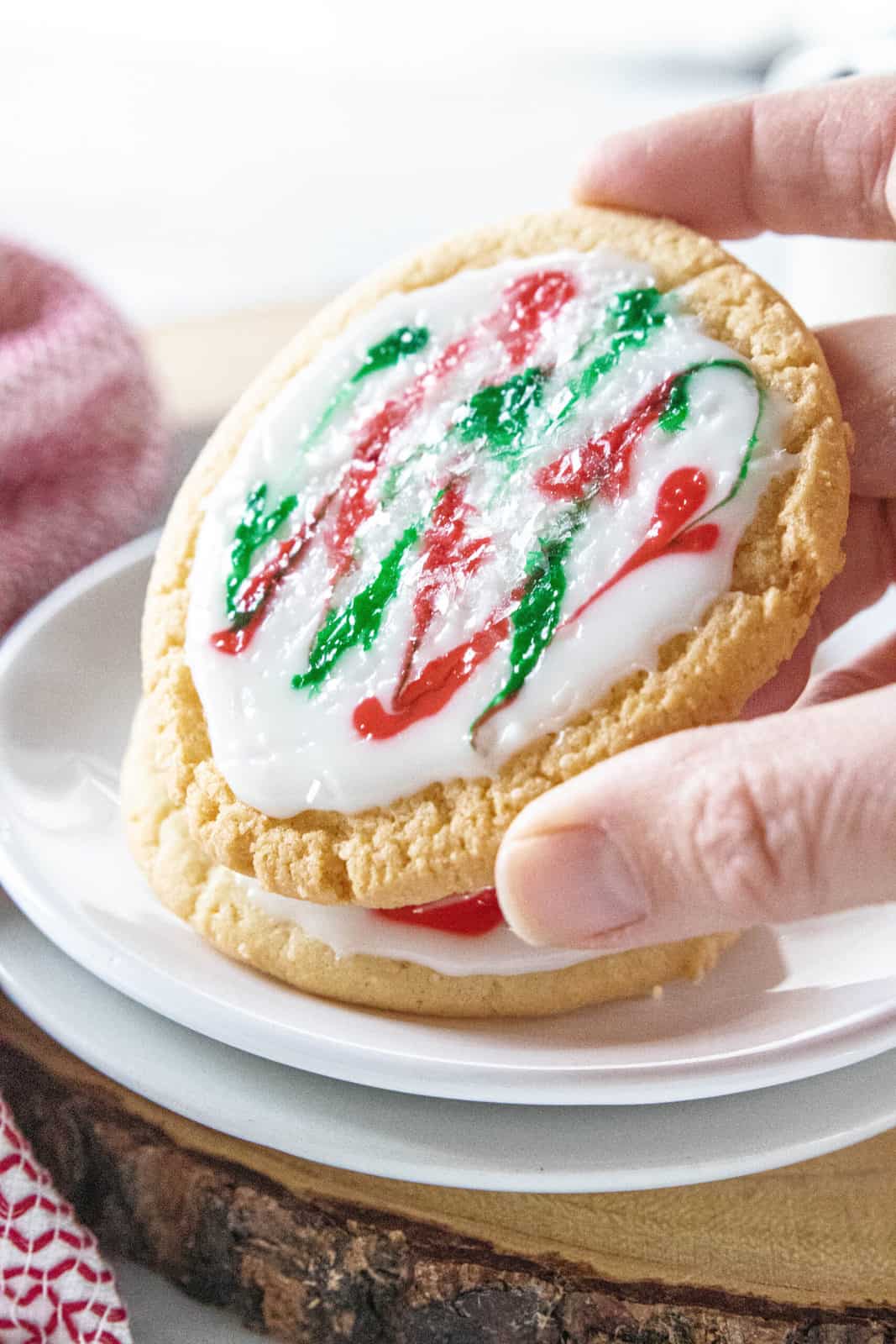 A hand picking up a frosted sugar cookie from a white plate with a stack of cookies. 