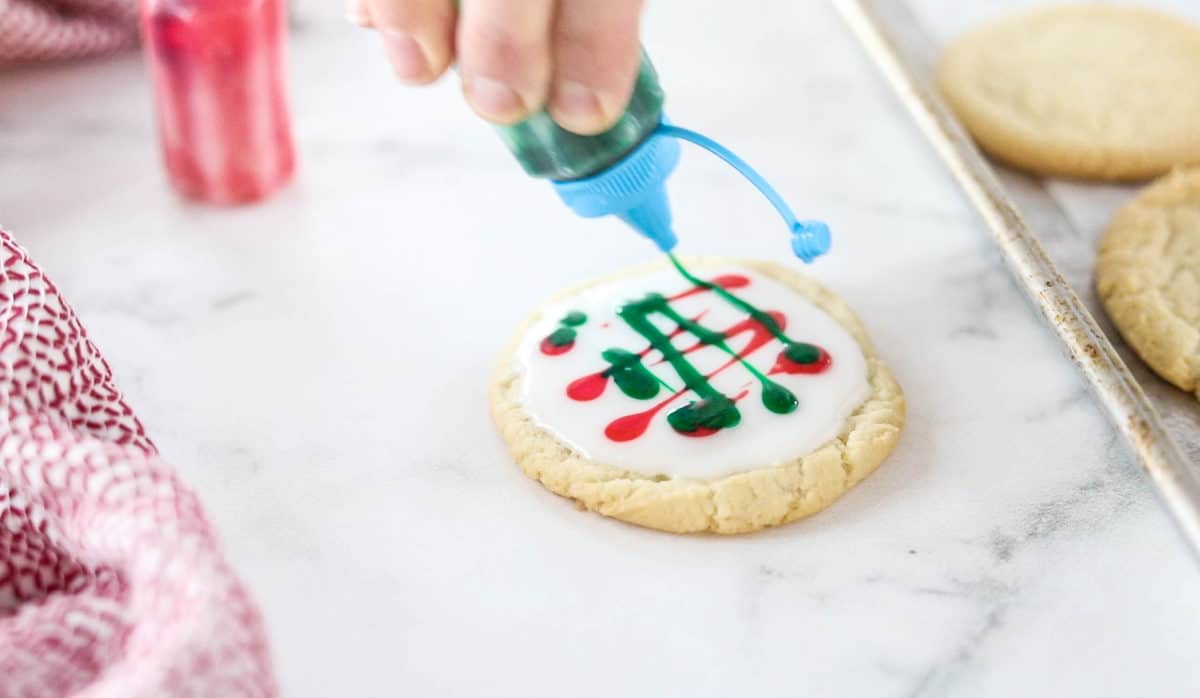 A hand adding green icing to a white frosted sugar cookie with red swirls. 