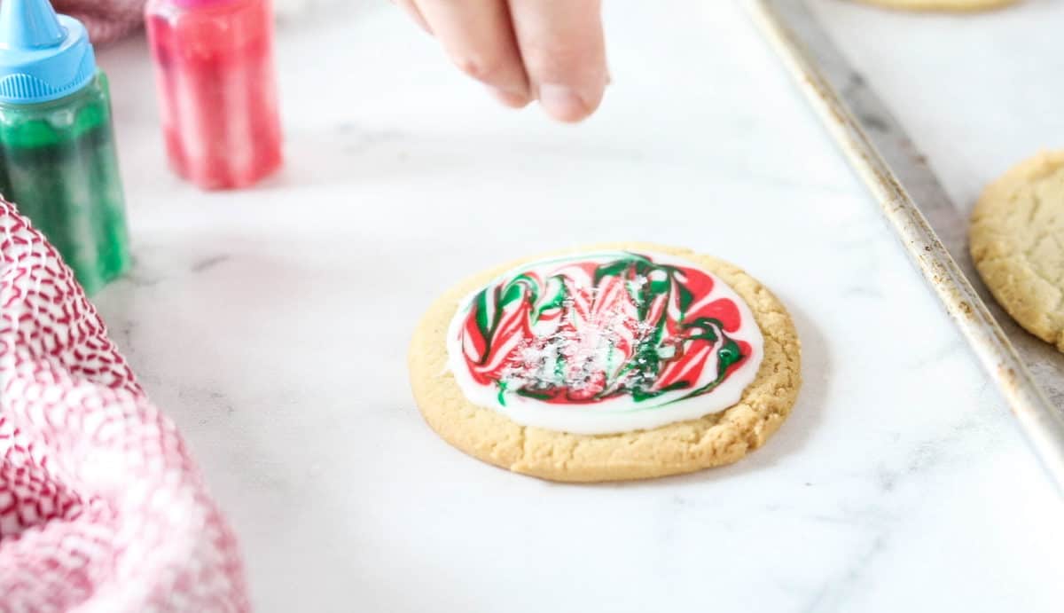 A hand being shown sprinkling sprinkles on top of a frosted Christmas cookie. 
