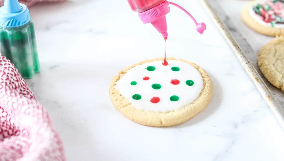 A white frosted sugar cookie being shown on a counter top with red and green icing dots on top of the white icing. 