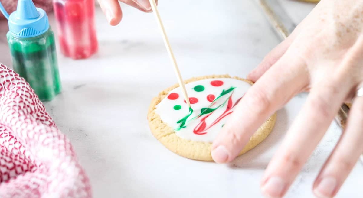 A hand using a toothpick to gently drag dots of red and green icing together to blend into the white icing. 