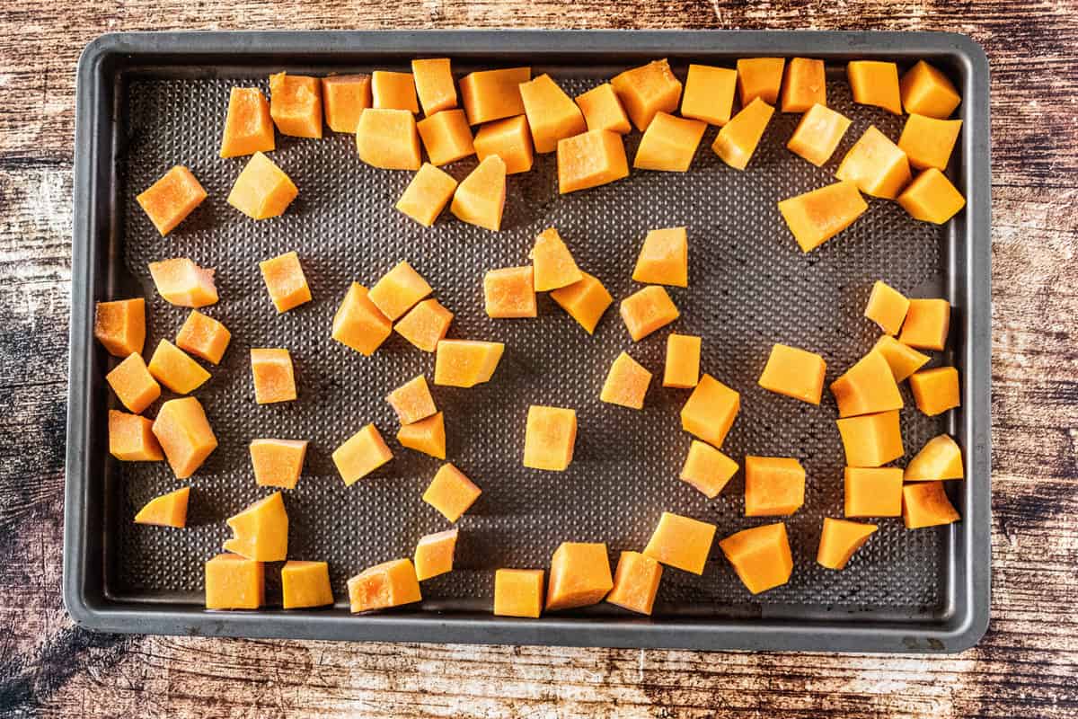 Butternut squash that has been chopped and added to a baking sheet that is on top of a wooden countertop. 