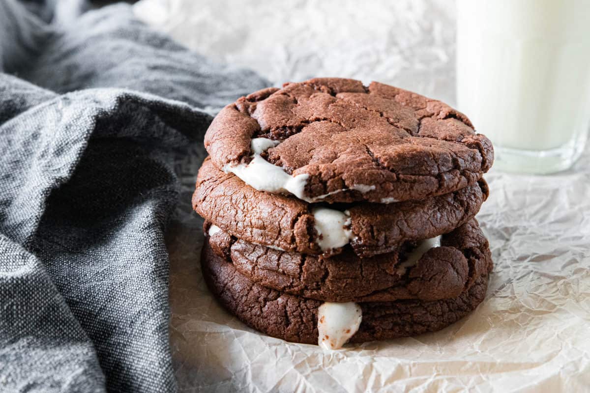 A stack of chocolate marshmallow cookies being shown on top of a piece of parchment paper with a dish towel next to the cookies. 