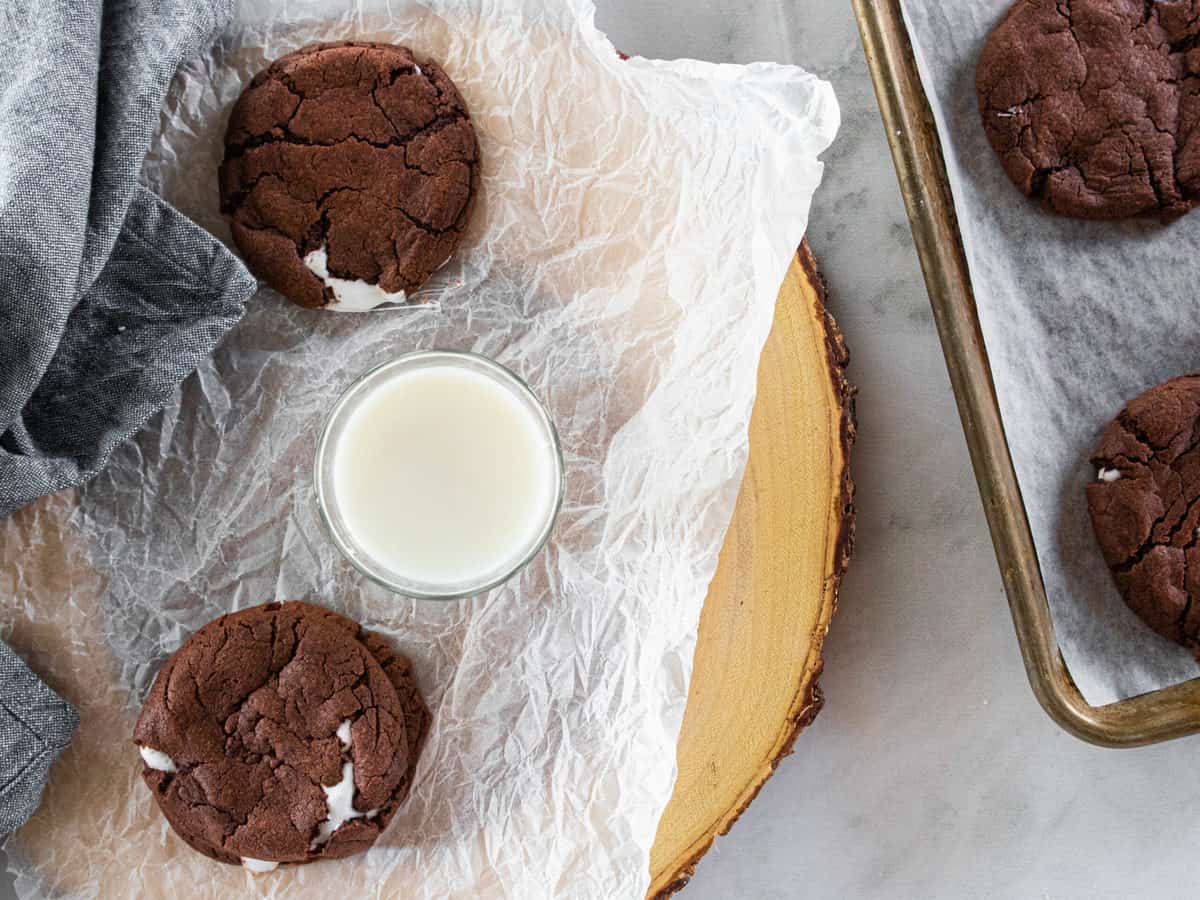 Cookies being shown on a piece of parchment paper with a glass of milk between two cookies. A baking sheet of cookies is next to the parchment paper. 