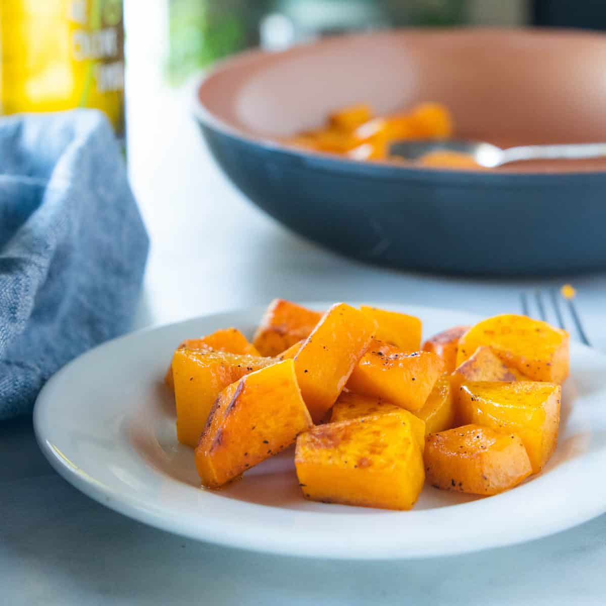 Sauteed butternut squash on a white plate with a fork next to the plate and a large serving bowl filled with sautéed squash behind the white plate.