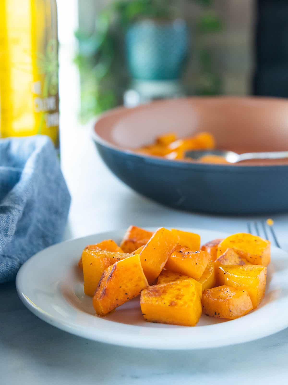 Sautéed butternut squash on a white plate with a fork next to the plate and a large serving bowl filled with sautéed squash behind the white plate.