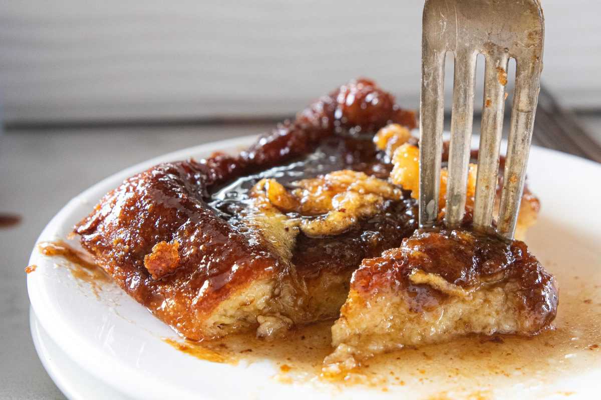 A fork being shown picking up a bite of French toast from a white plate. 