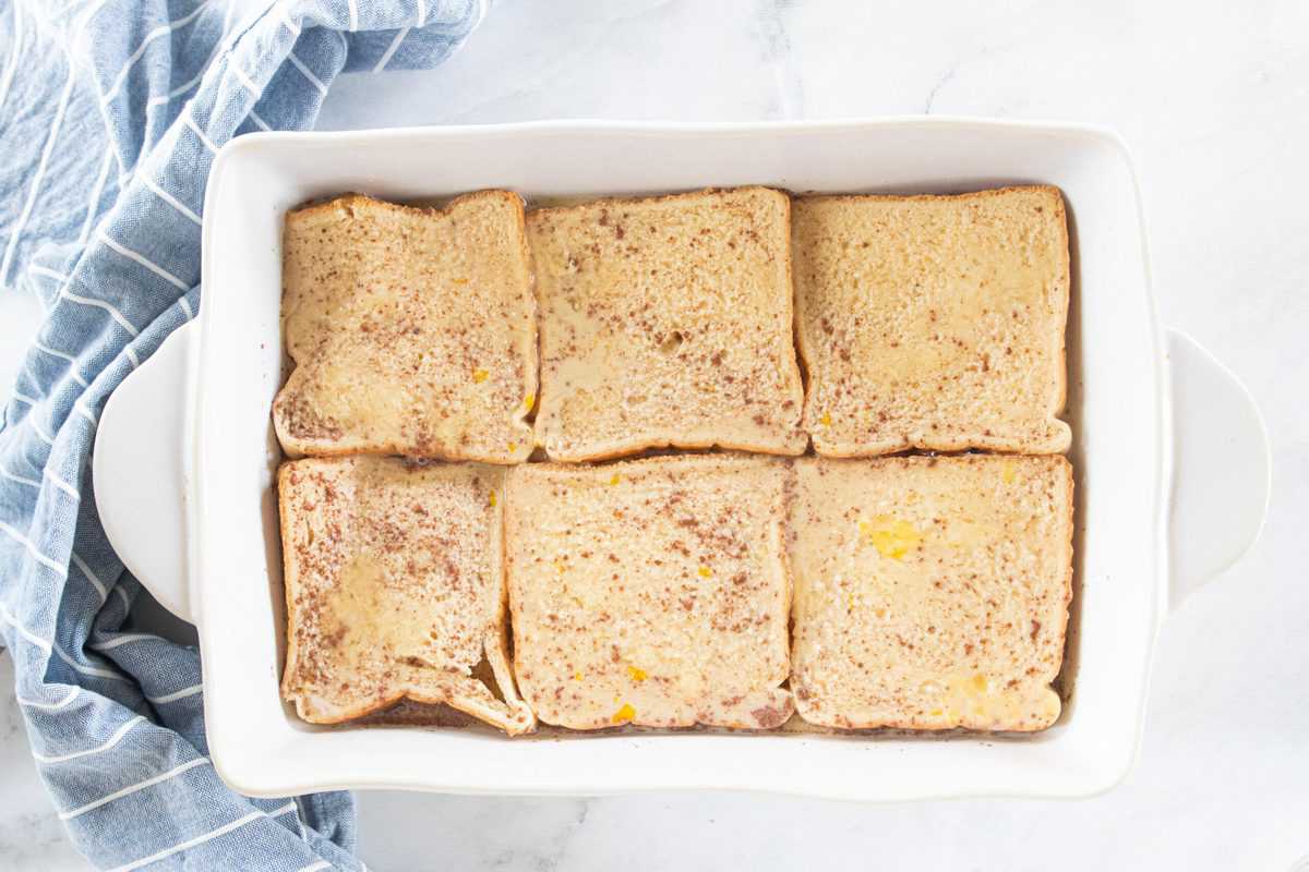 A white baking dish filled with slices of white bread covered with an egg mixture layered on top of a brown sugar mixture.