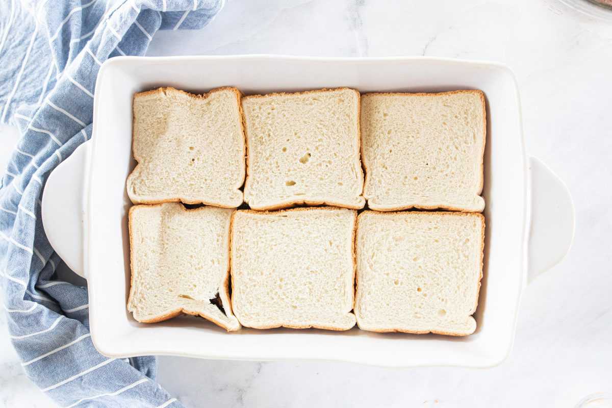 A white baking dish with slices of white bread layered on top of a brown sugar mixture being shown. 