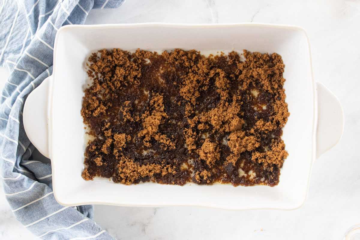 A white baking dish filled with a brown sugar mixture on top of a countertop with a blue dish towel next to the dish. 