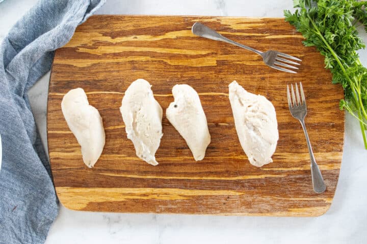 Four chicken breasts shown on a wooden cutting board with two forks on the board all on top of a countertop.