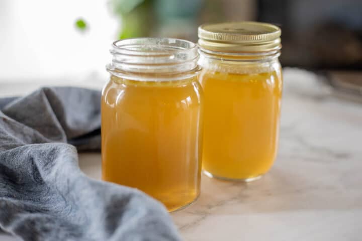 Chicken broth being shown in mason jars on top of a countertop with a blue dish towel next to the jars. 