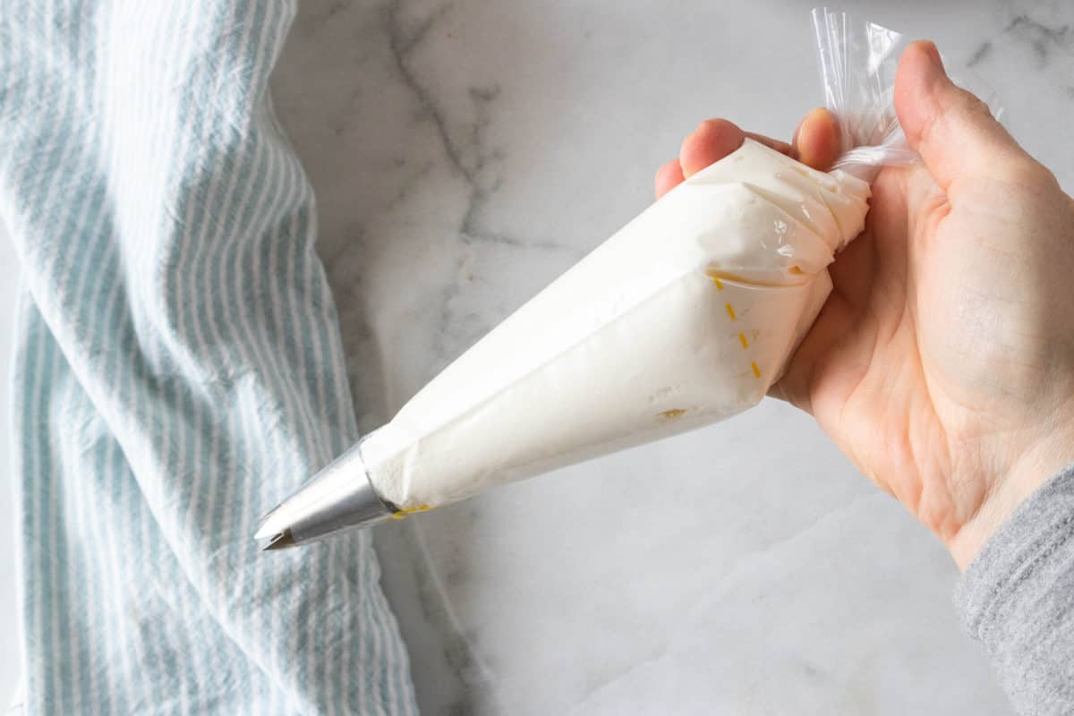 Almond buttercream frosting being shown in a decorating bag that is held in a hand over a countertop. 
