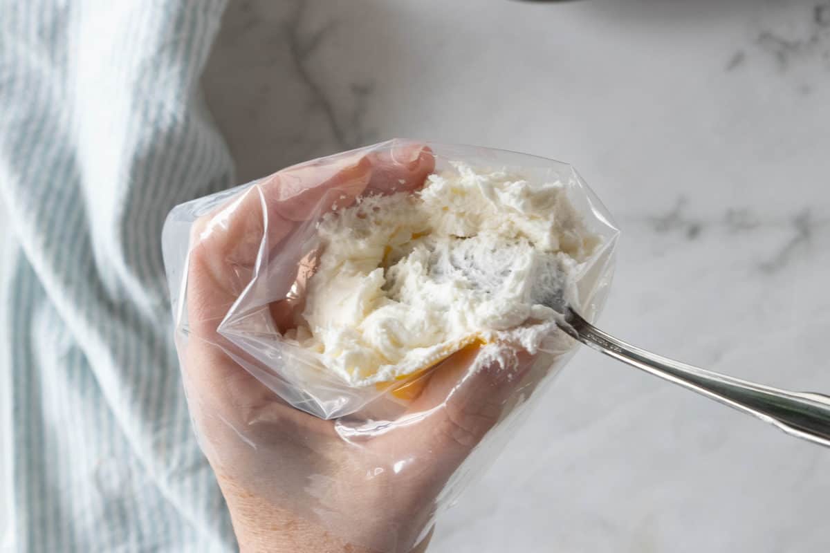 A hand holding a decorating bag with that is being filled by a spoon with almond buttercream frosting.