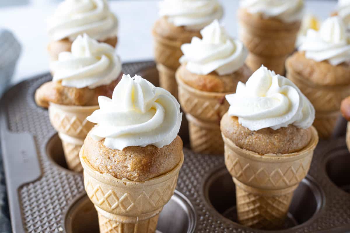 A muffin tin being shown filled with ice cream cones that are stuffed with cake and topped with buttercream frosting. 