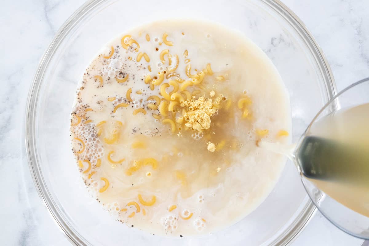 Ingredients for crockpot Mac and cheese being shown in a glass mixing bowl with chicken broth being poured into the bowl.
