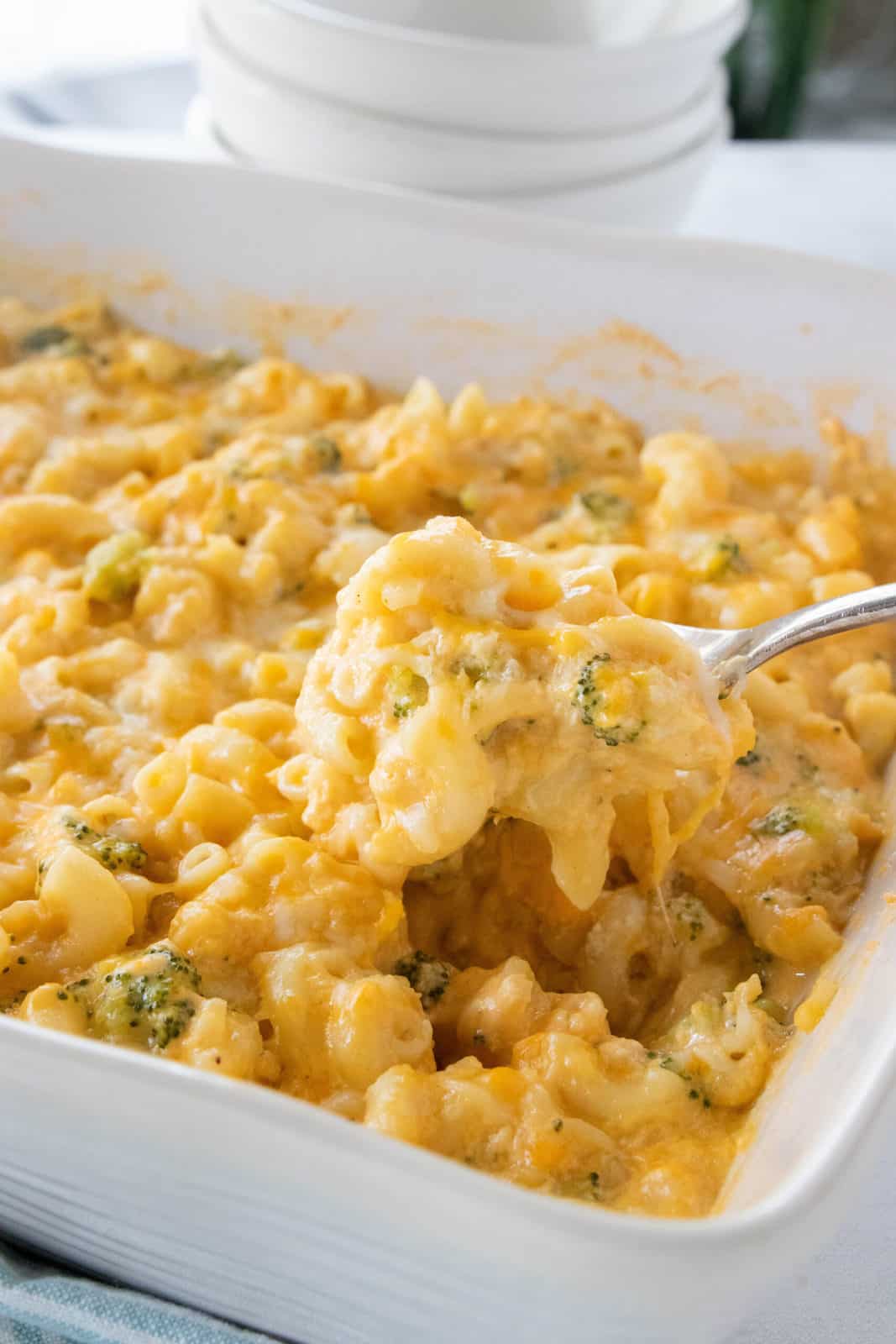 Mac and cheese with veggies being shown in a white baking dish with a spoon scooping a serving from the dish.