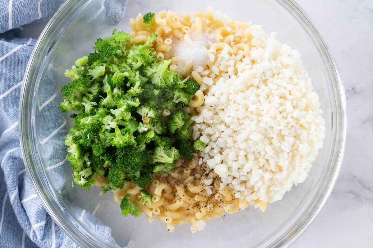 Broccoli, riced cauliflower, elbow pasta, salt and pepper being shown in a large glass mixing bowl that is on top of a granite countertop. 
