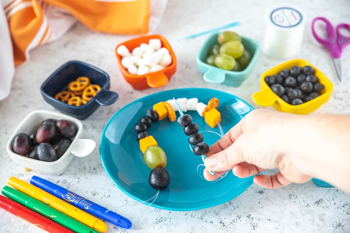 A snack necklace with blueberries being threaded onto the string. 