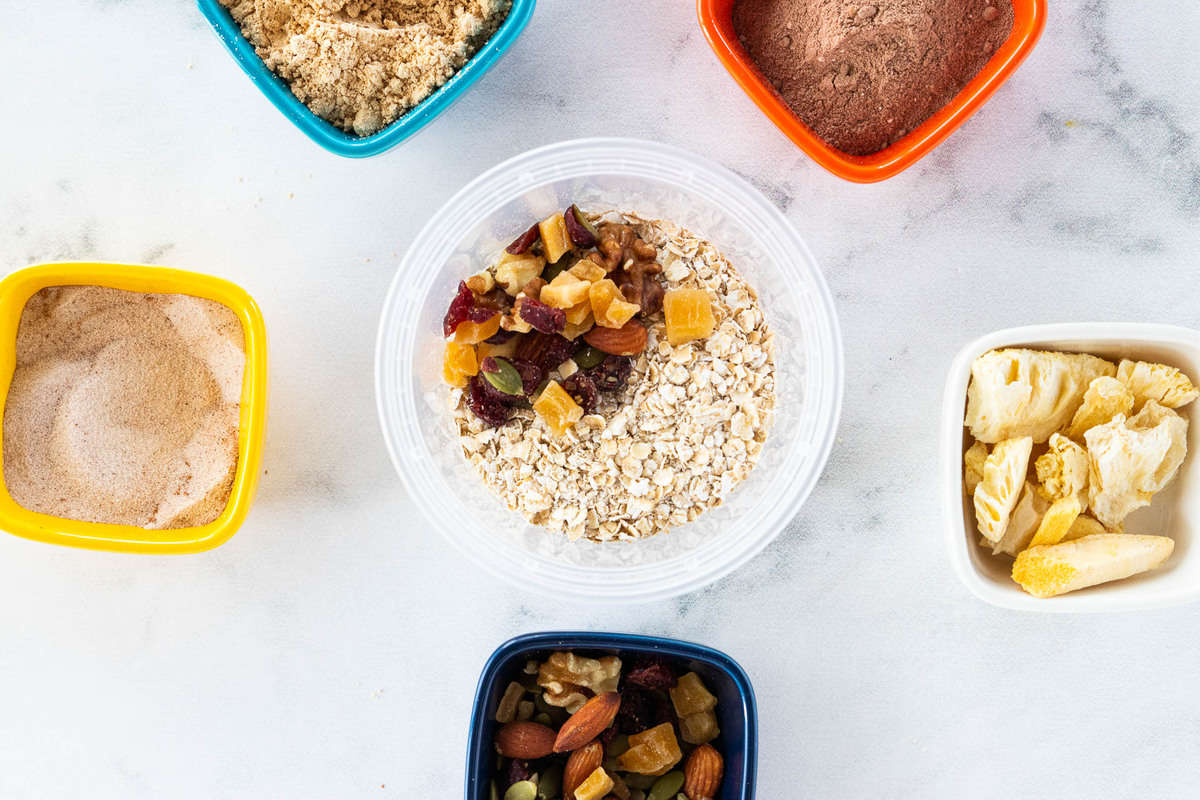 A container of oats with trail mix on top surrounded by a variety of toppings 